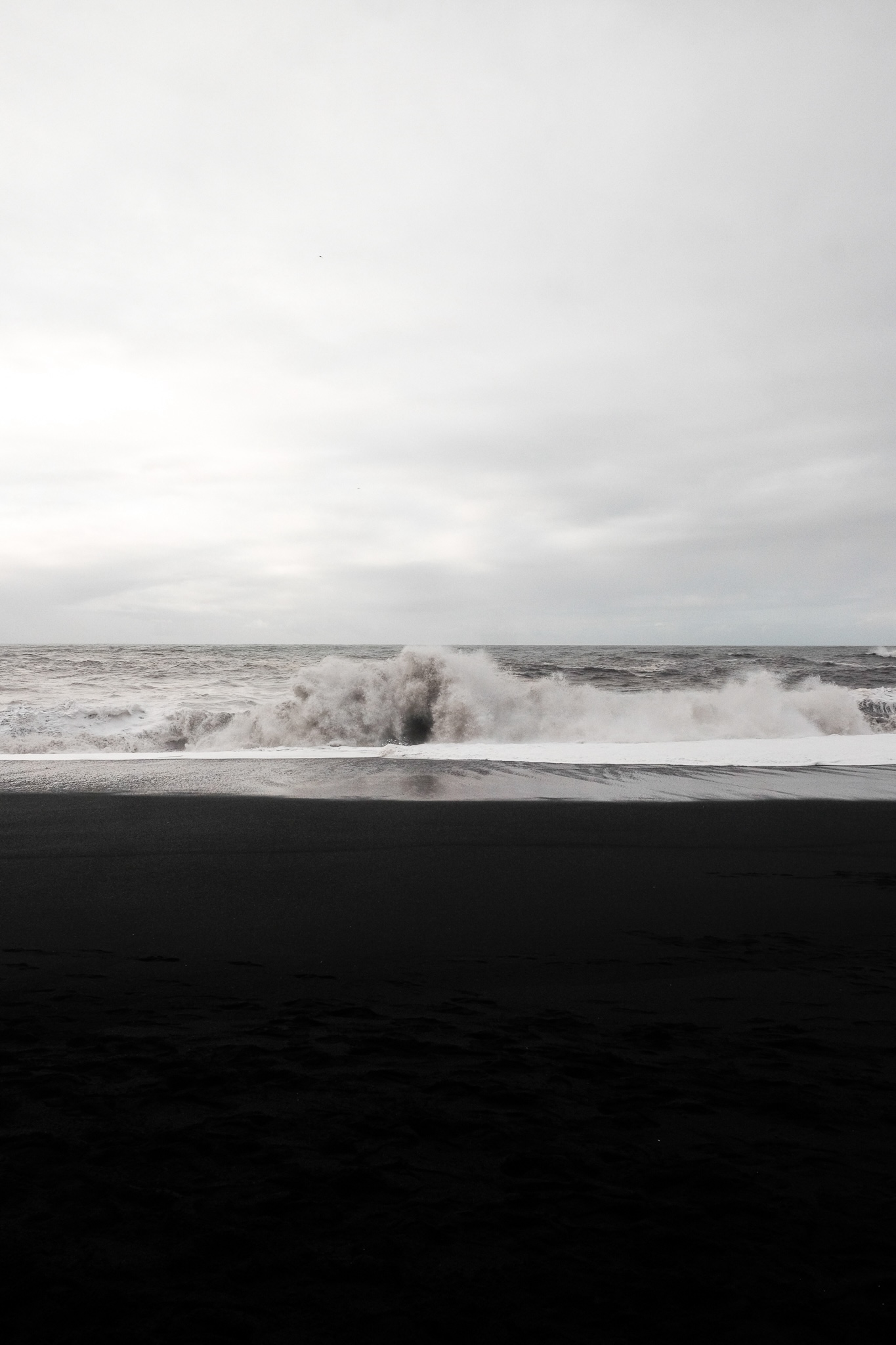 Slightly brown sea wave crashes on the black beach, the sky white and overcast, the water cuts the black half of the image in two sections of colour creating a visually pleasing pattern