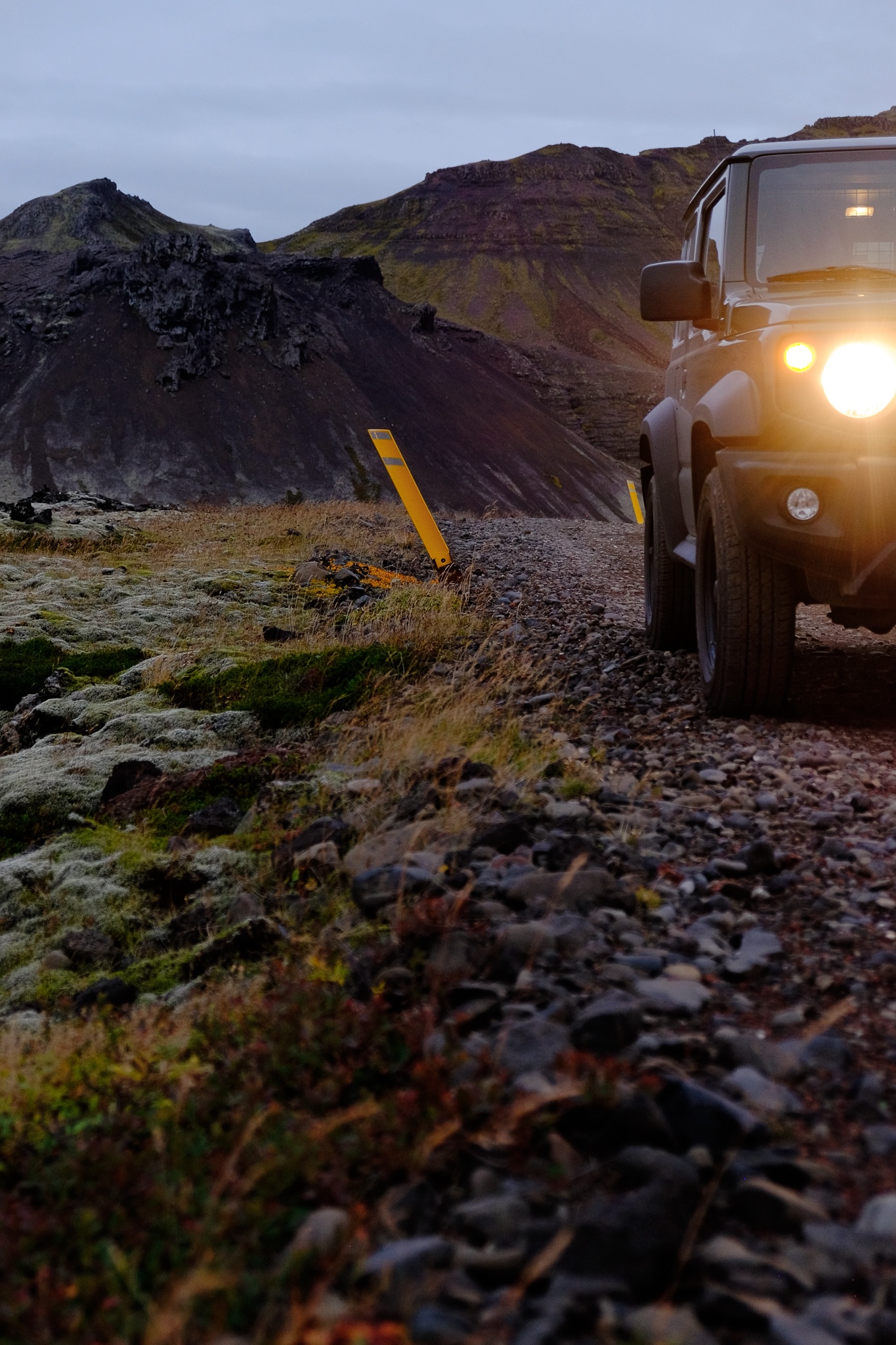 A small deep army green 4x4 with warm lights face you on a gravel road next to moss covered volcanic rock and a mountain in the background