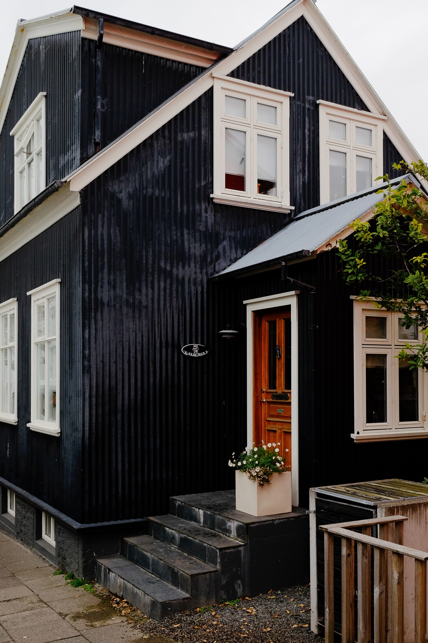 A black corrugated house with white window frames and a wooden door has a cute square pot with flowers in it