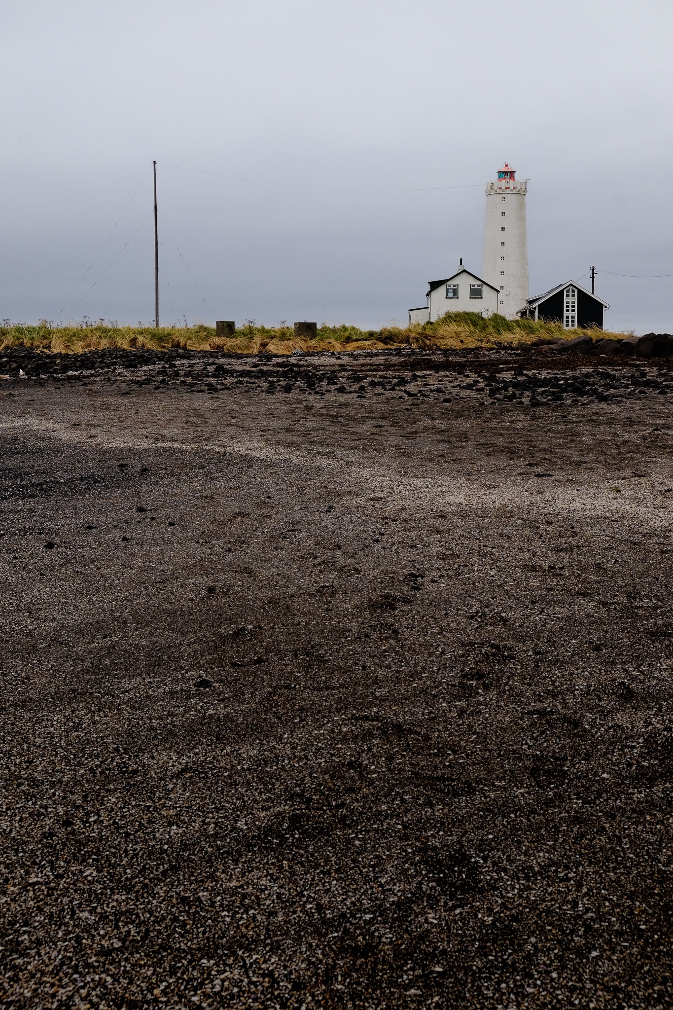 A beach of black sand and white shells with grass and a lighthouse in the distance