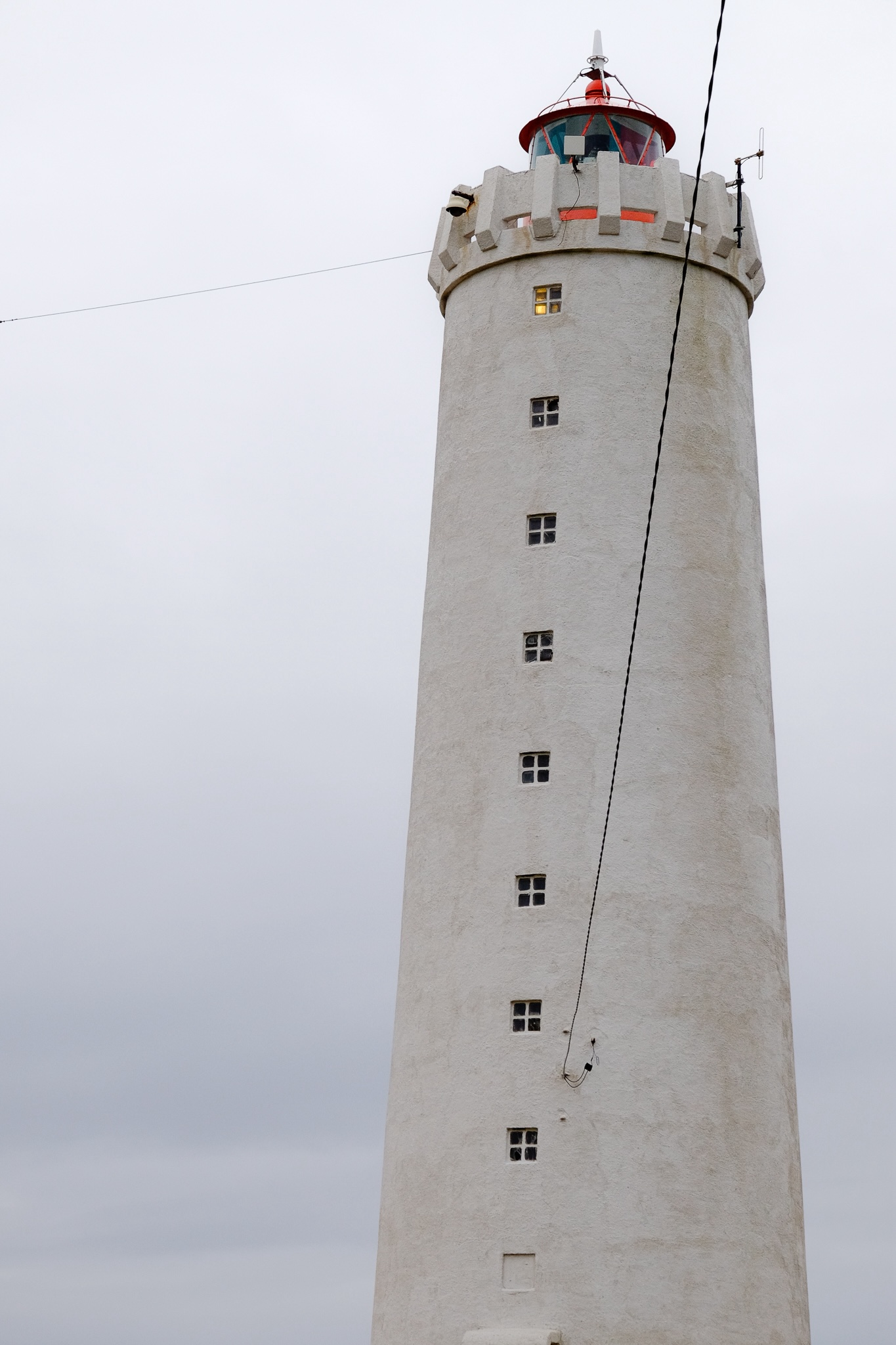 A weathered white lighthouse with red housing for the massive bulb with cables extending from two parts of the structure