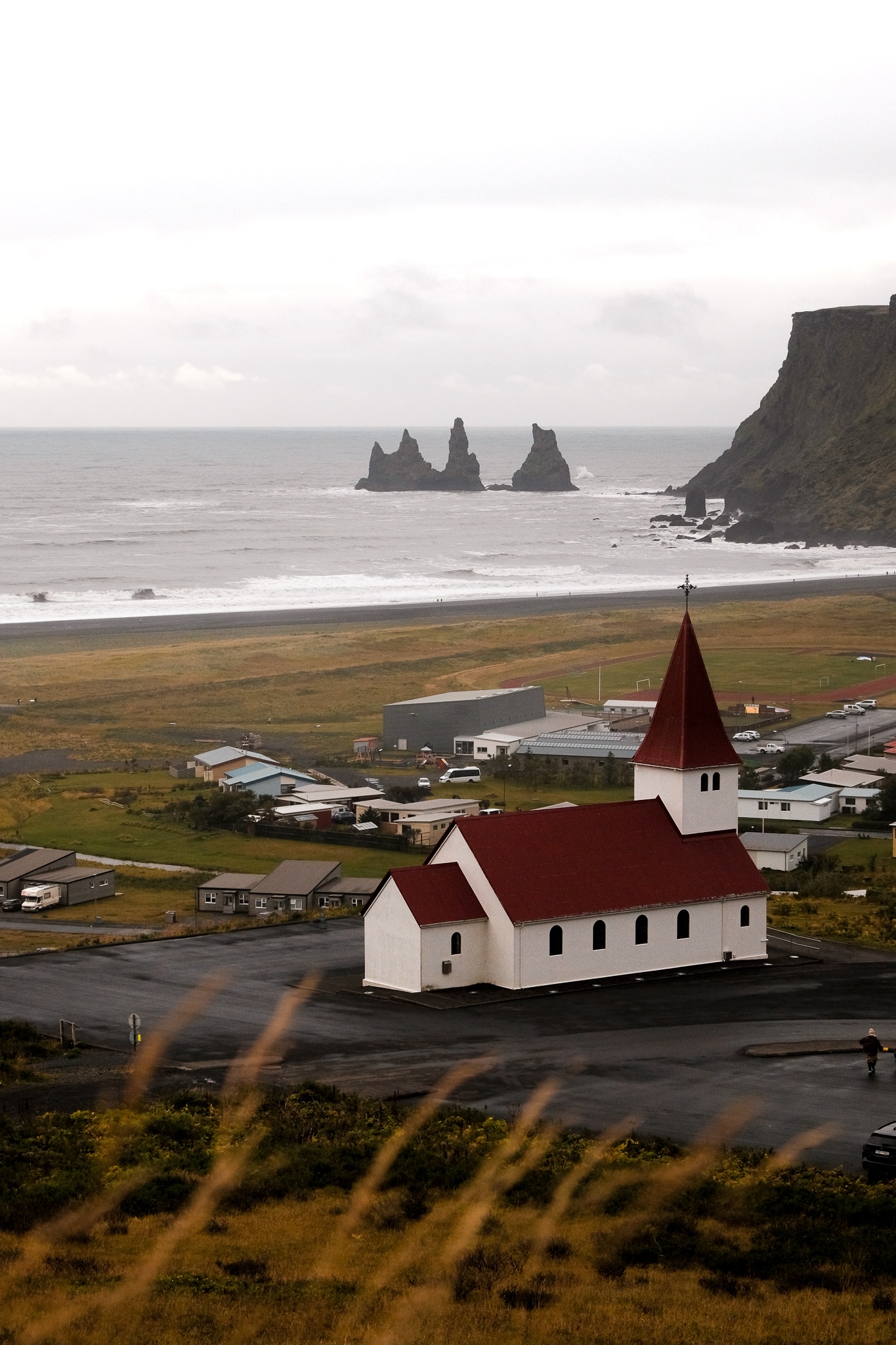 Vik from the mountain side showing off it's beautiful white church with a red roof, the black beach in the distance and yellow grass blowing in the wind in the foreground