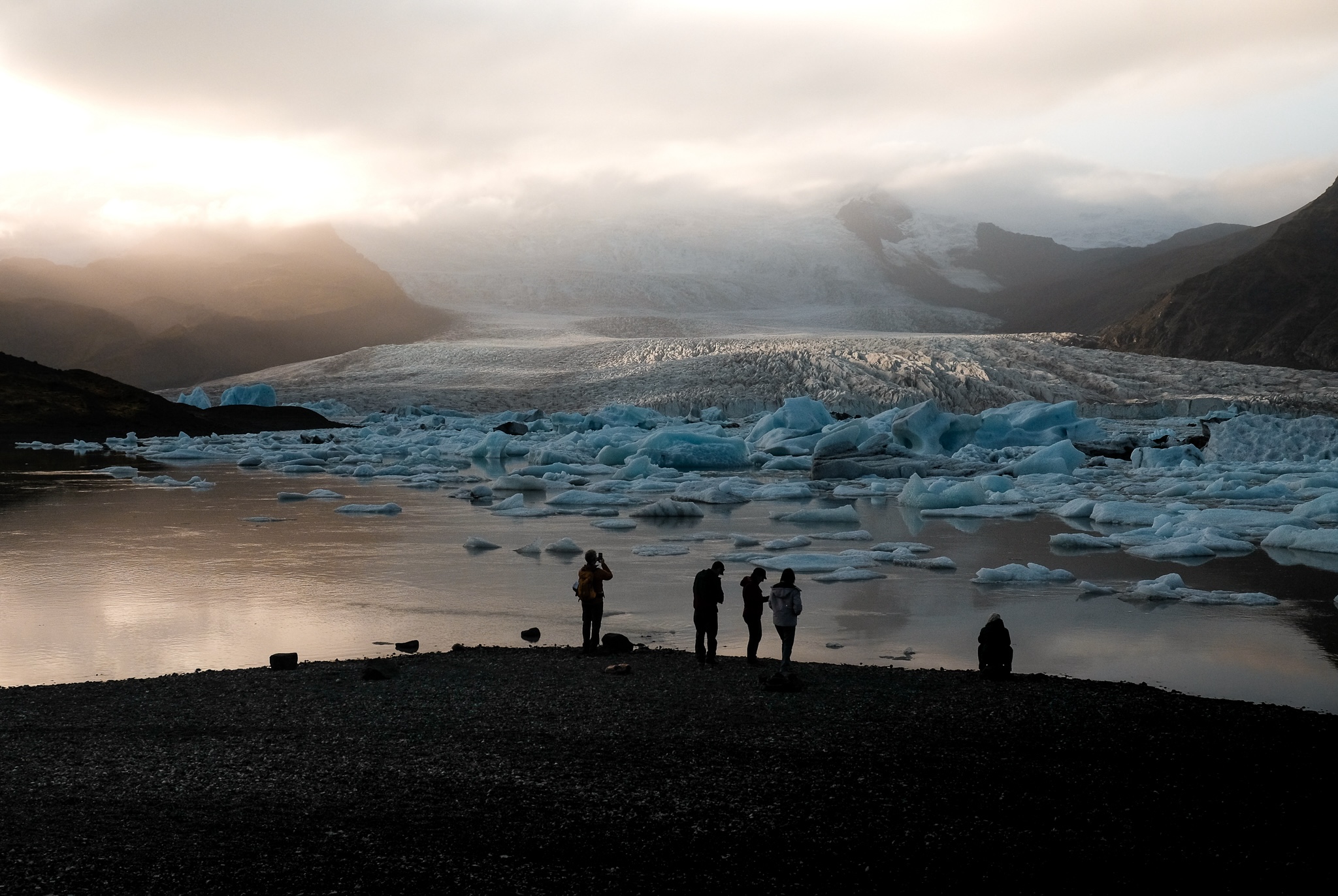 A silhouette of people enjoying the view and photographing a glacier moving down a mountain into a glaciel lake with large chunks of floating ice
