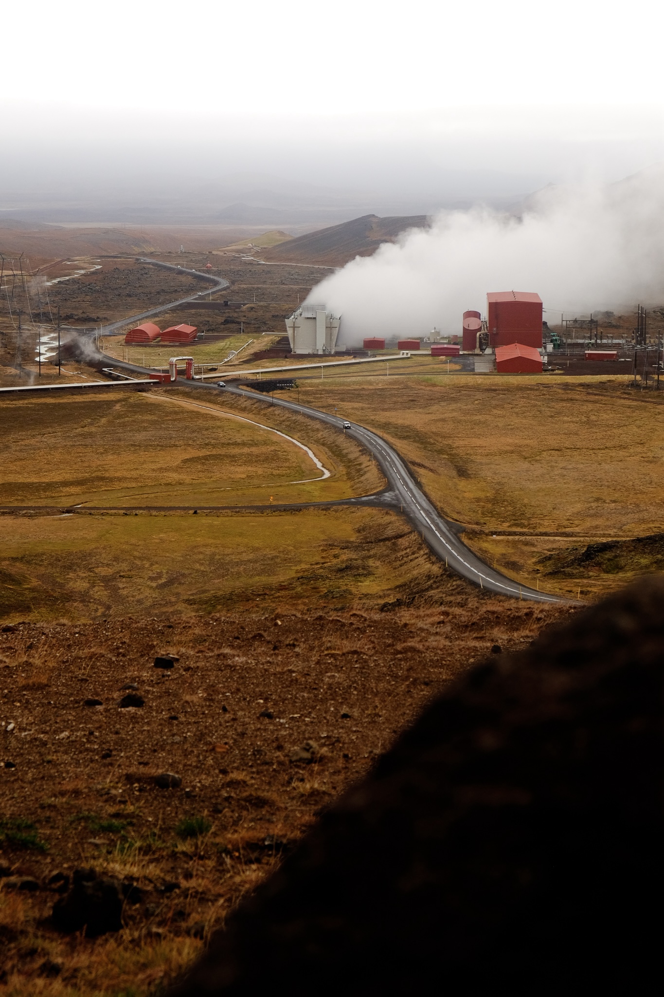 A geothermal powerplant of red buildings is gushing with white steam against a brown and yellow landscape