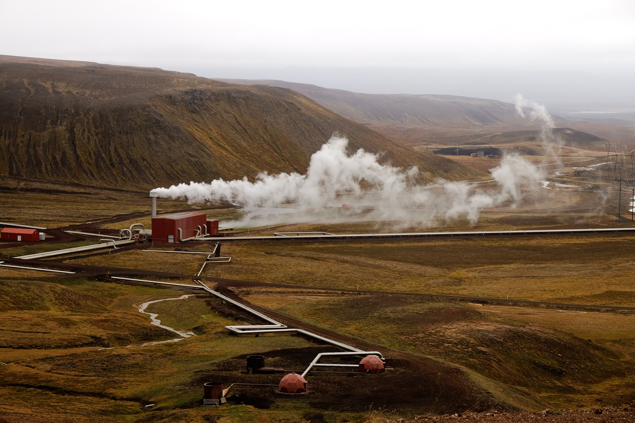 A geothermal powerplant red building is gushing white steam from a vent, the steam travels across the landscape as it dissapates into the air