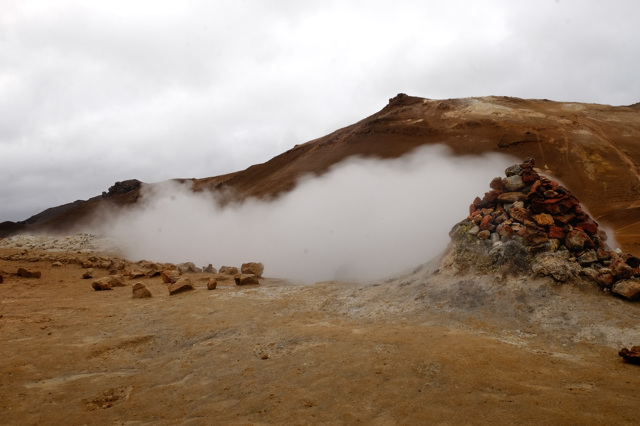 Geothermal steam geiser of red and brown rock billows with white steam and travels across a yellow-brown barren landscape