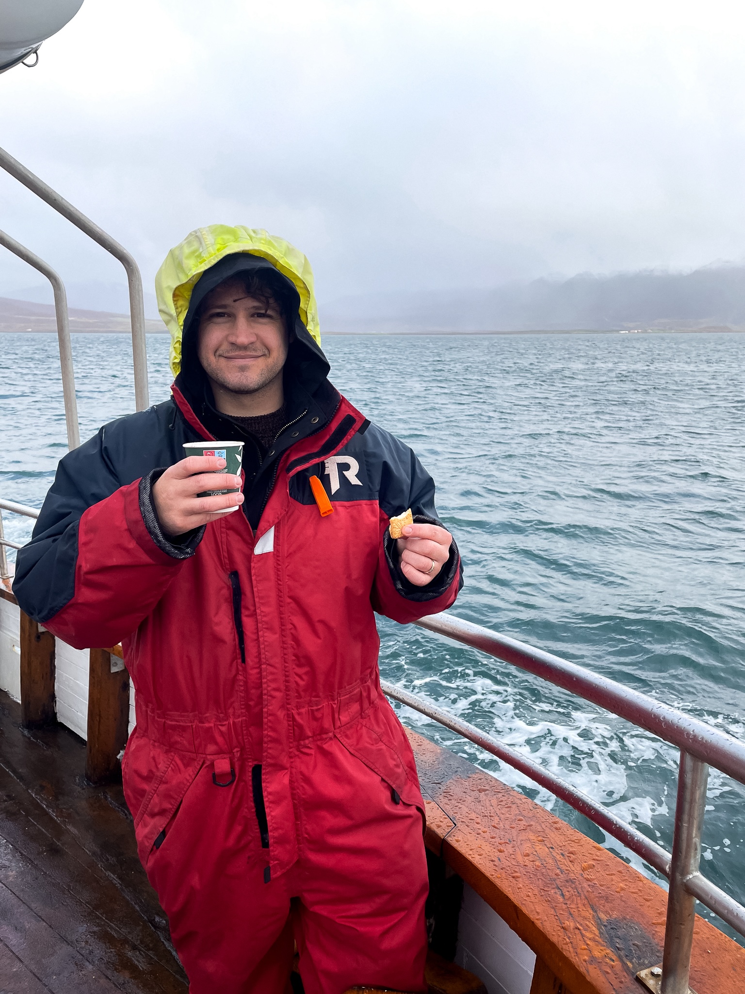 Simon with a red waterproof suit holding hot chocolate and a cheap biscuit smiles with the water of the fjord and mountains in the backgroun