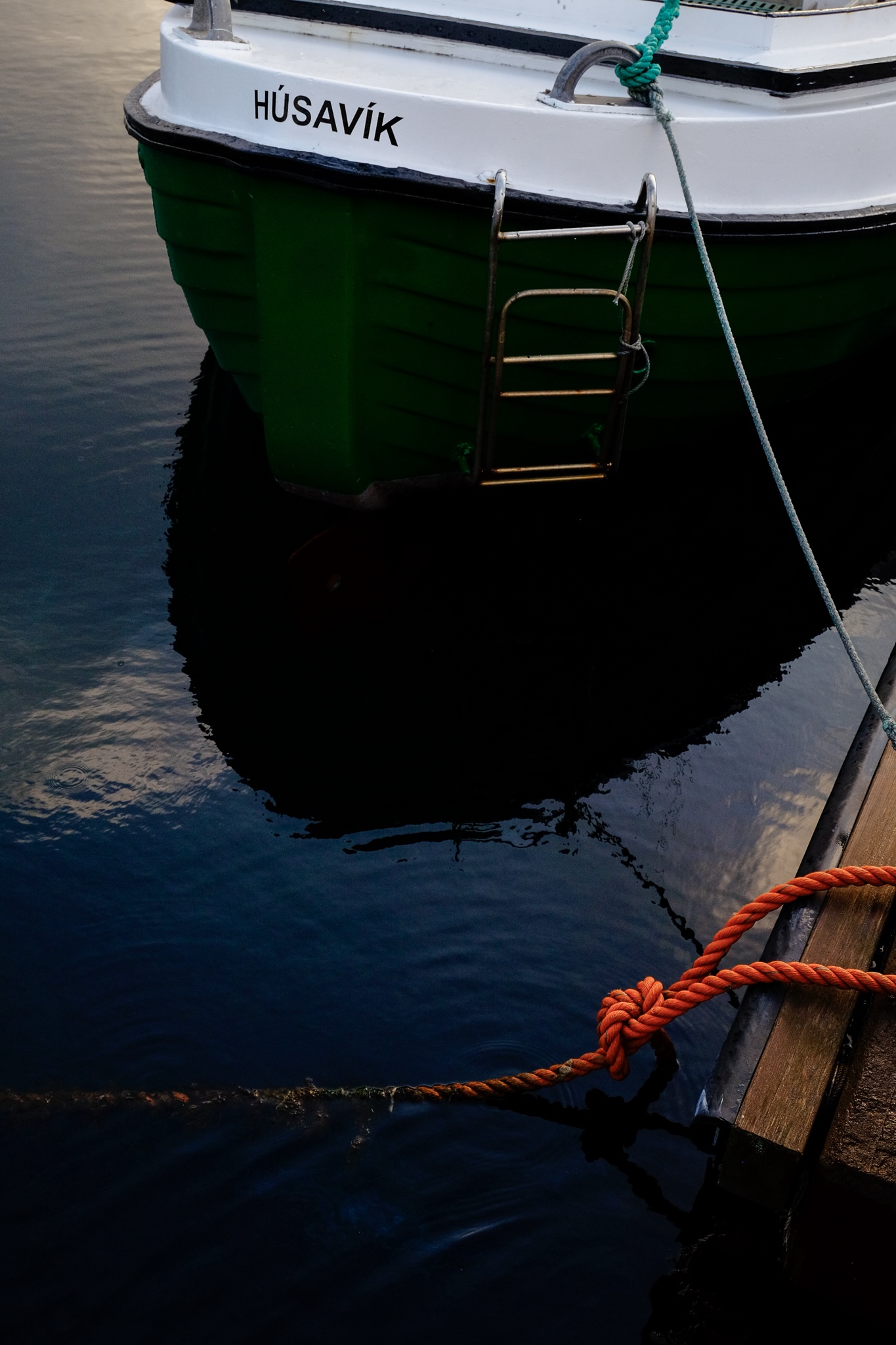 Deep green boat named Husavik against dark blue water in the harbour with bright orange rope holding another boat moored to the dock
