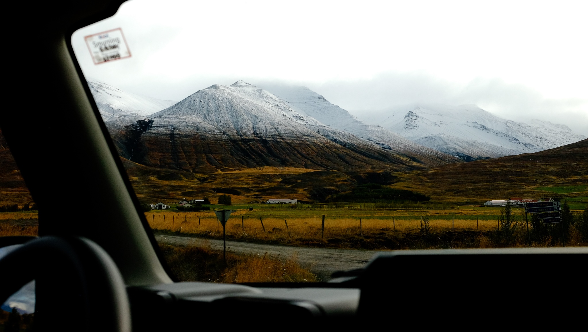 From the window of a car with the dashboard in the foreground you see a yellow landscape transition to a snowy mountain fade into a white sky