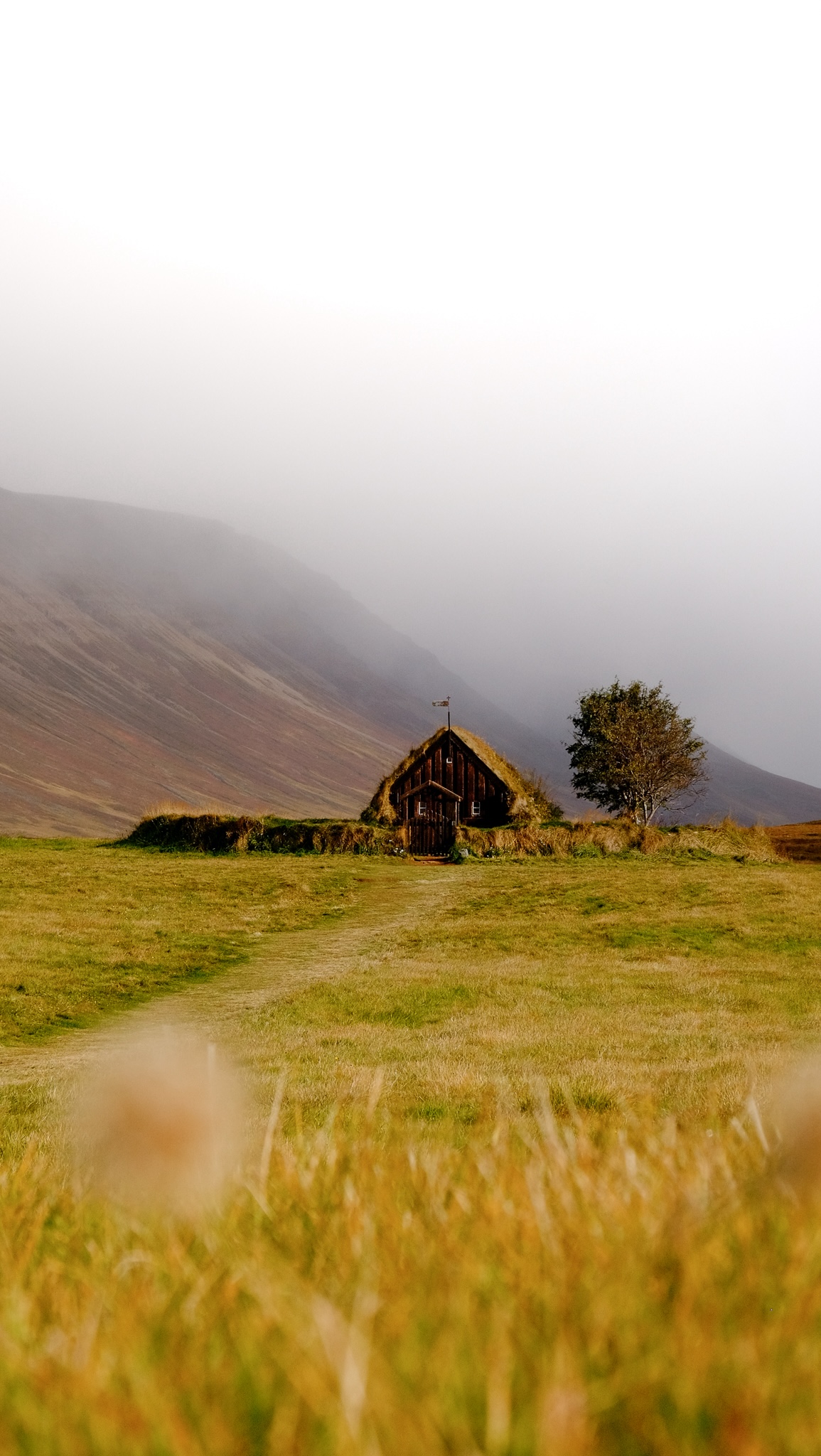 An old church with a grass roof and circular enclosure in the distance from a grass field with a mountain in the background with misty clouds rolling in