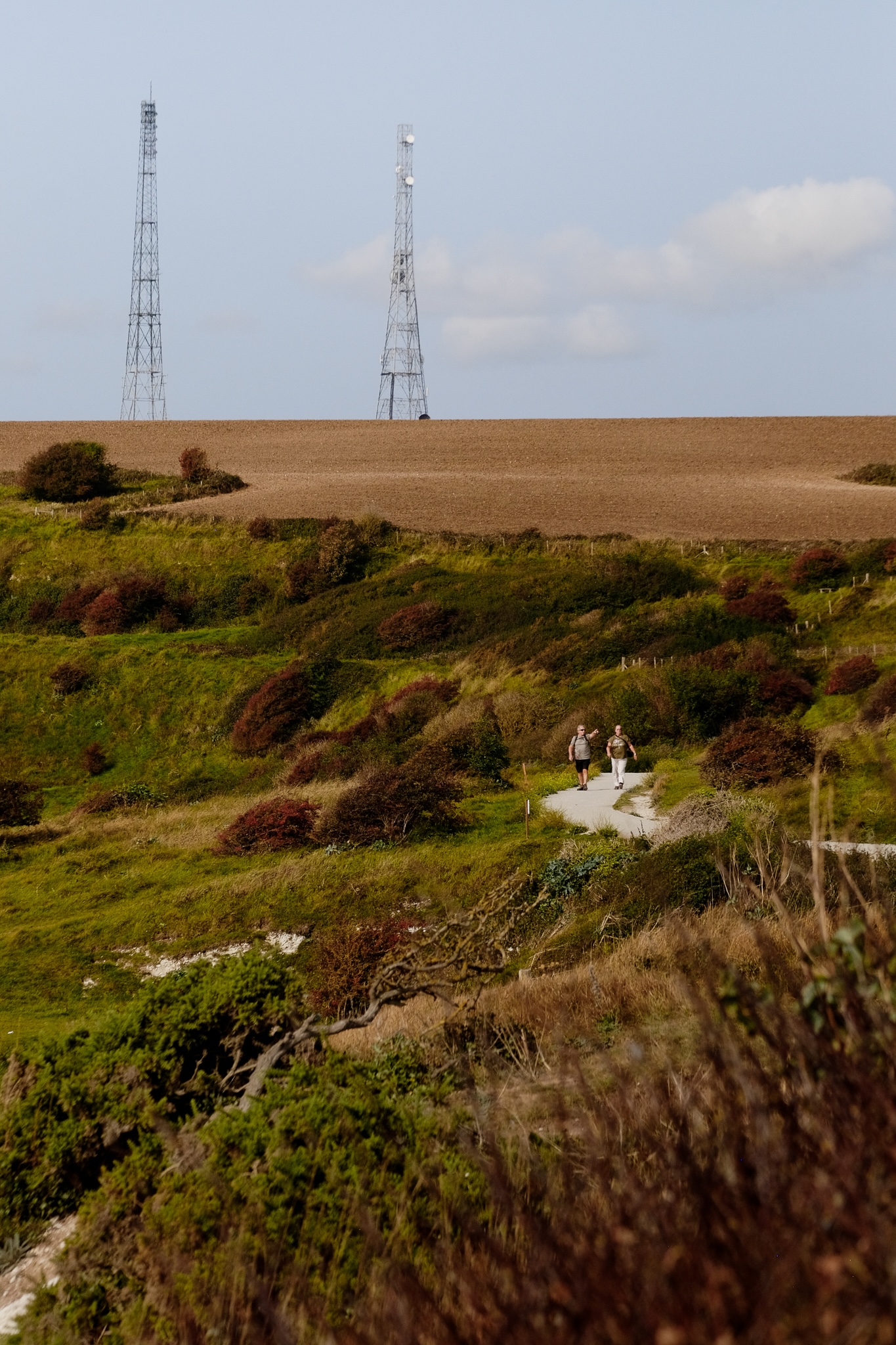 Two people walking down a path along the cliffs of Dover with farm land and cell towers in the background