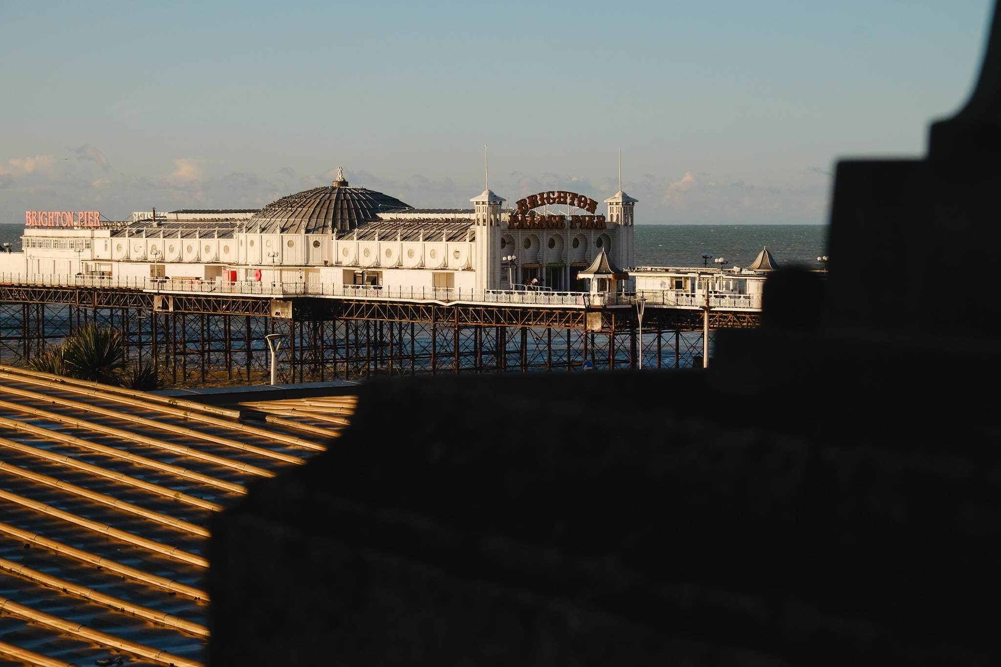 Brighton pier from the road peaks out from a roof in the foreground, the early morning sun casting a bright warm light on the structure