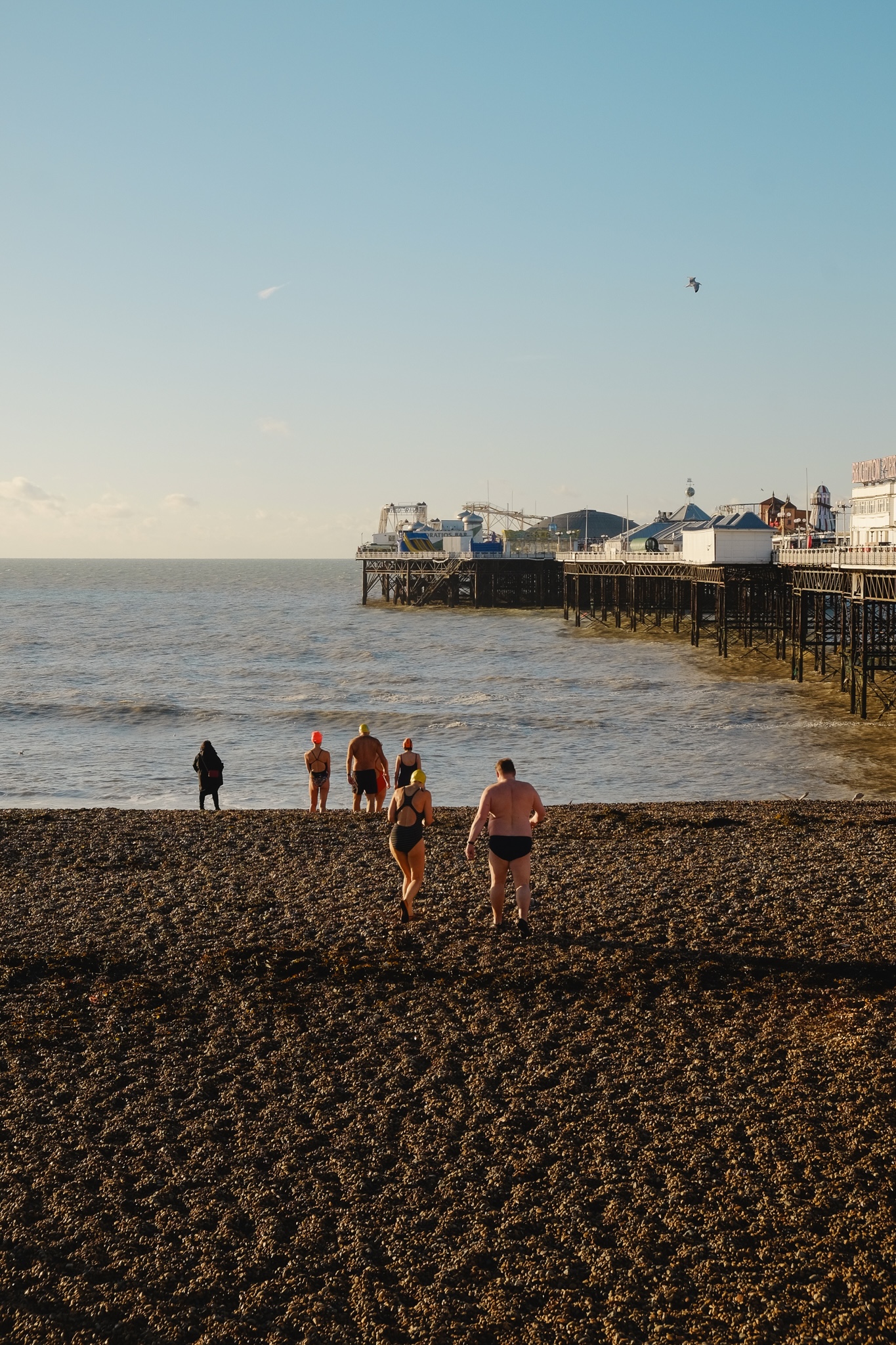 A group of swimmers make their way to the sea with the pier to the right, another person in a warm coat is standing looking into the distance