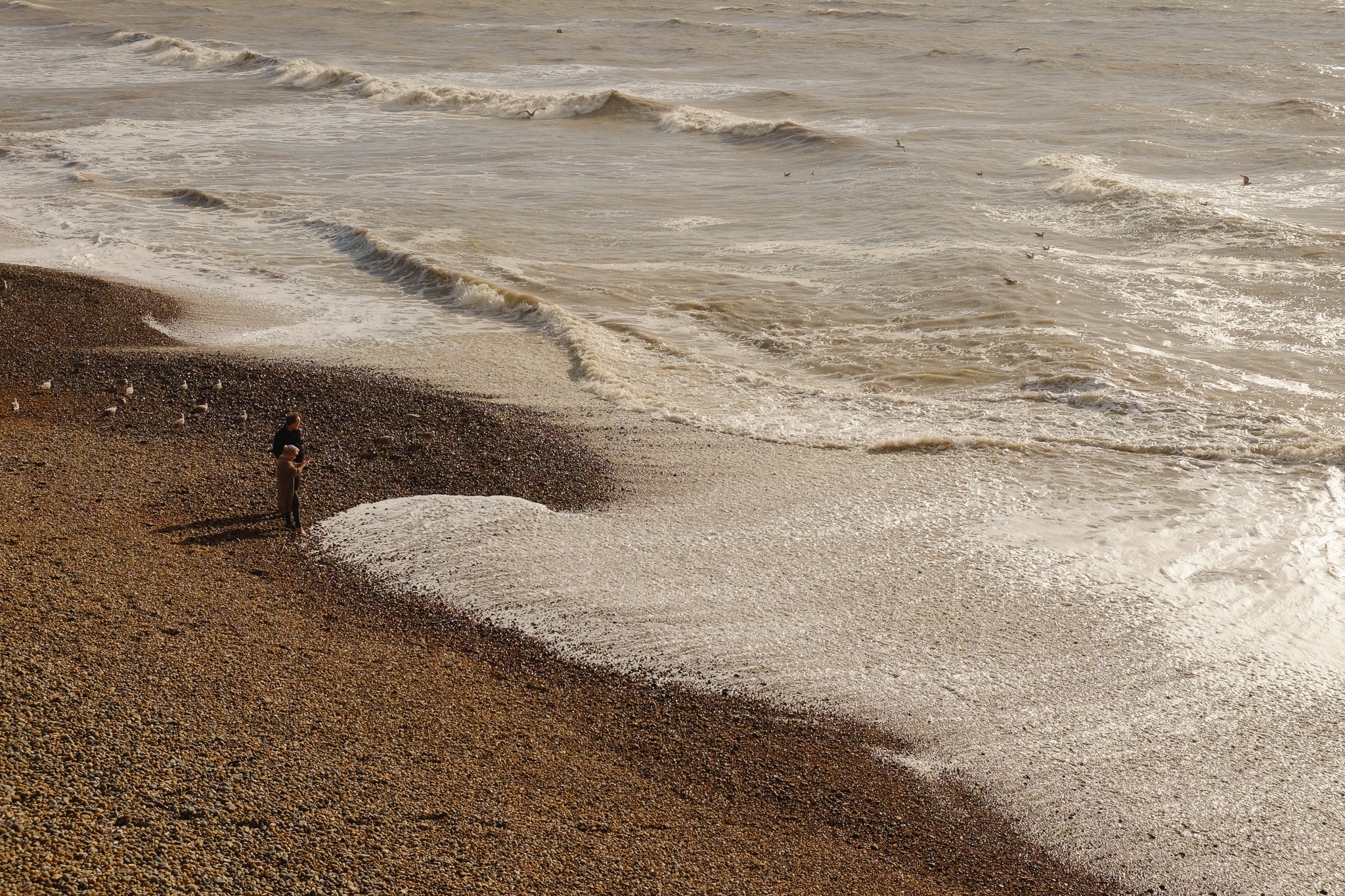 A couple stands on the pebbled beach photographing the waves as they crash up towards them and withdraw. The pebbles swell with sound as they bump each other, disturbed by the wave.