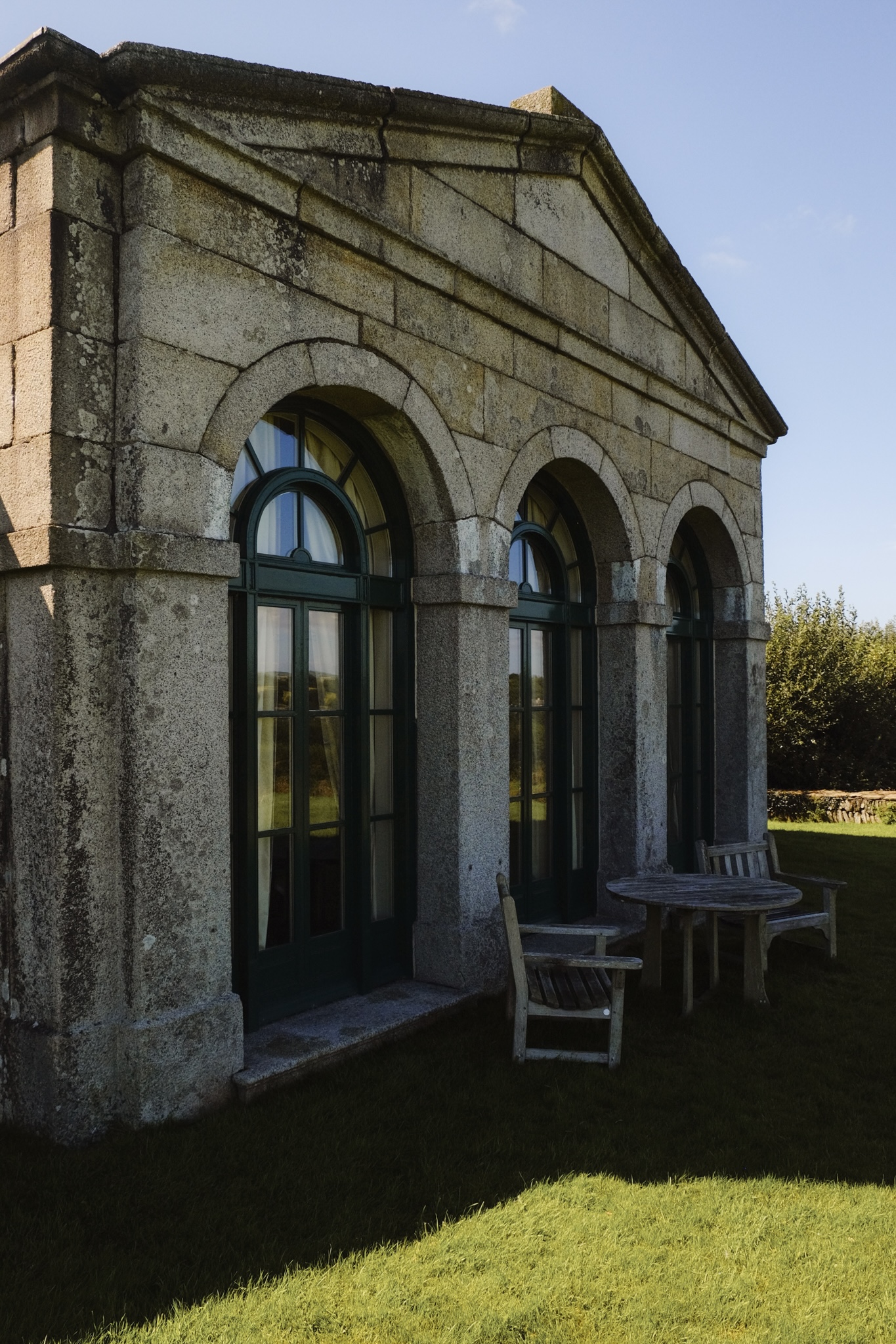 Front of an old stone building with a wooden table and chairs on the grass outside